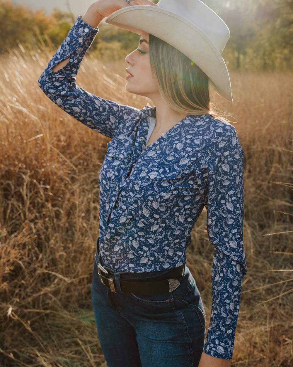 Mujer con sombrero blanco y camisa vaquera azul estampada, en un campo de pasto seco al atardecer