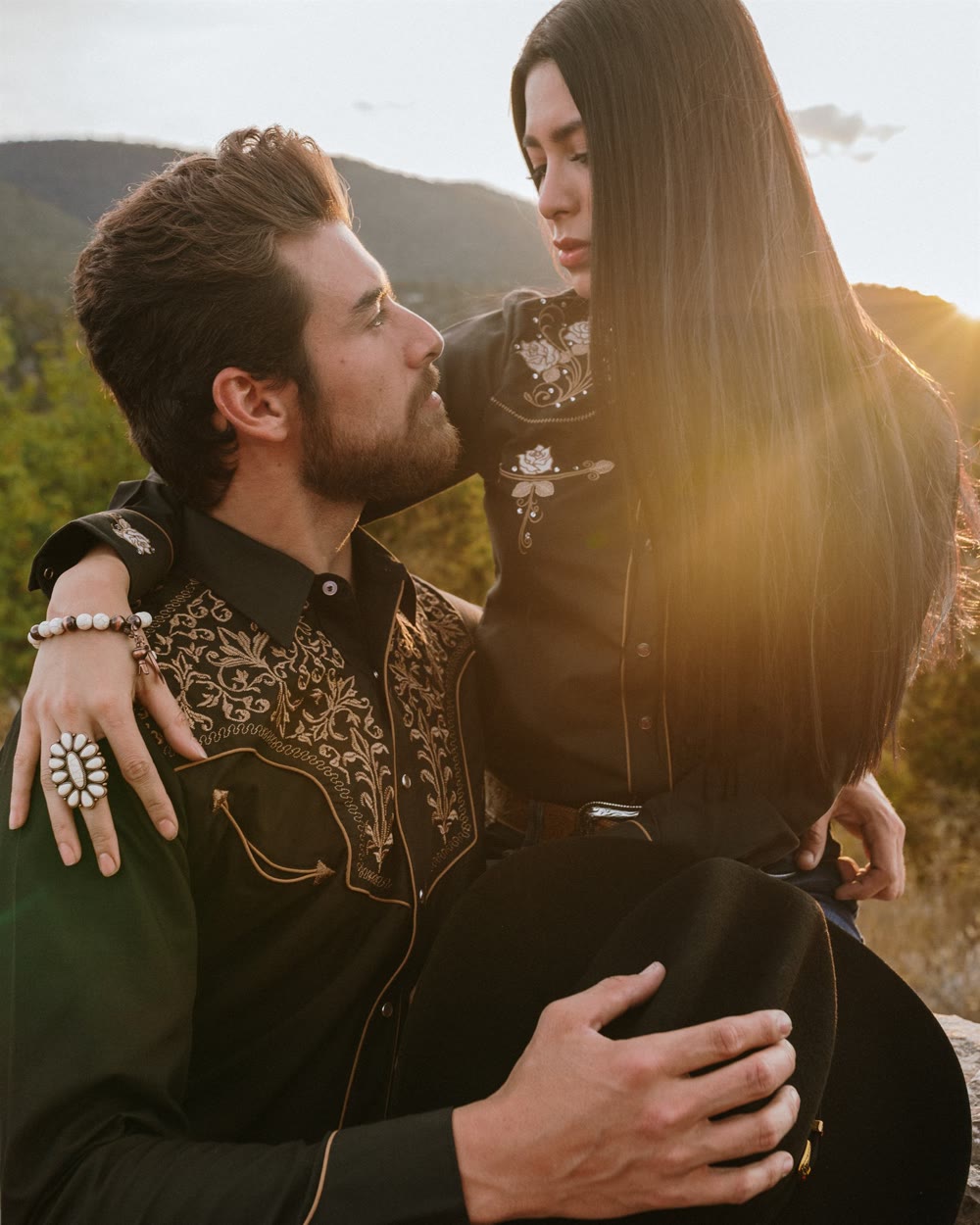 Pareja con camisas vaqueras bordadas se mira a contraluz, con la sierra al atardecer detrás